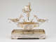 Profile view of elaborate silver double-oval centerpiece with swirling arms extending to two shallow side-dishes, with central fountain-like stem topped by classical figurines, all on a rectangular tray pedestal.  