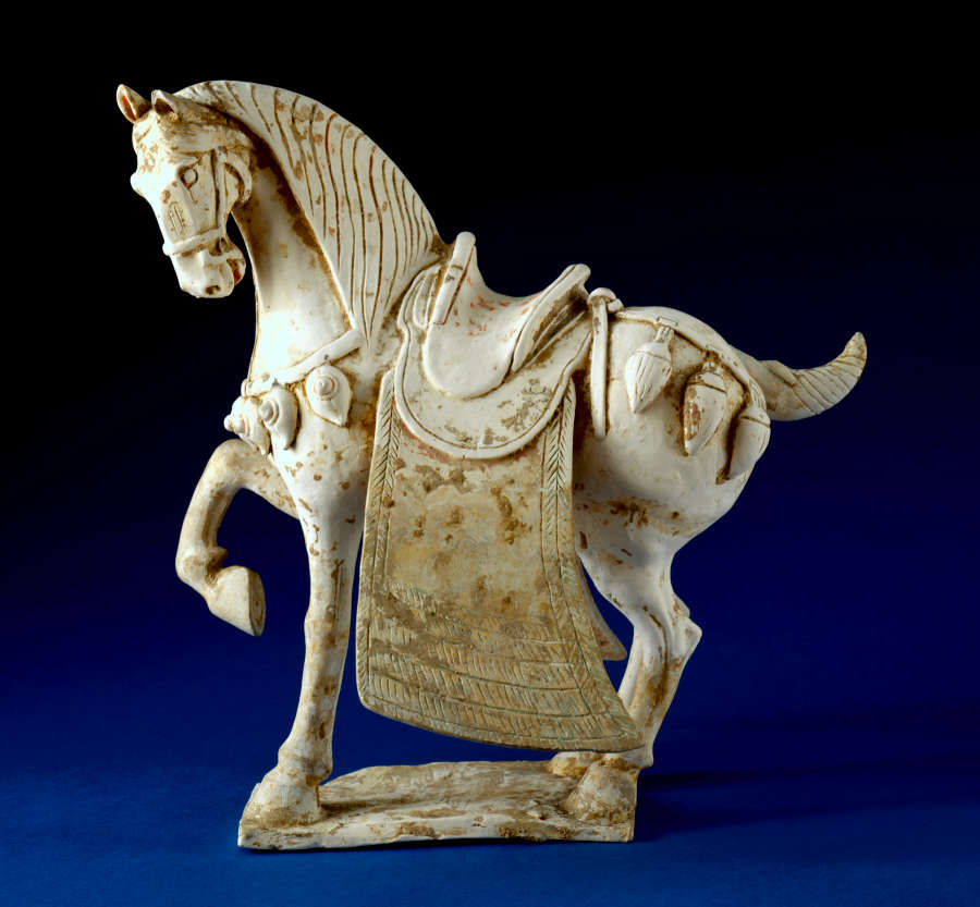 Side-view of a white ceramic sculpture of a saddled and ornamented horse with one foot raised, a short wound tail and heavy mane. It stands on a thin connected base.