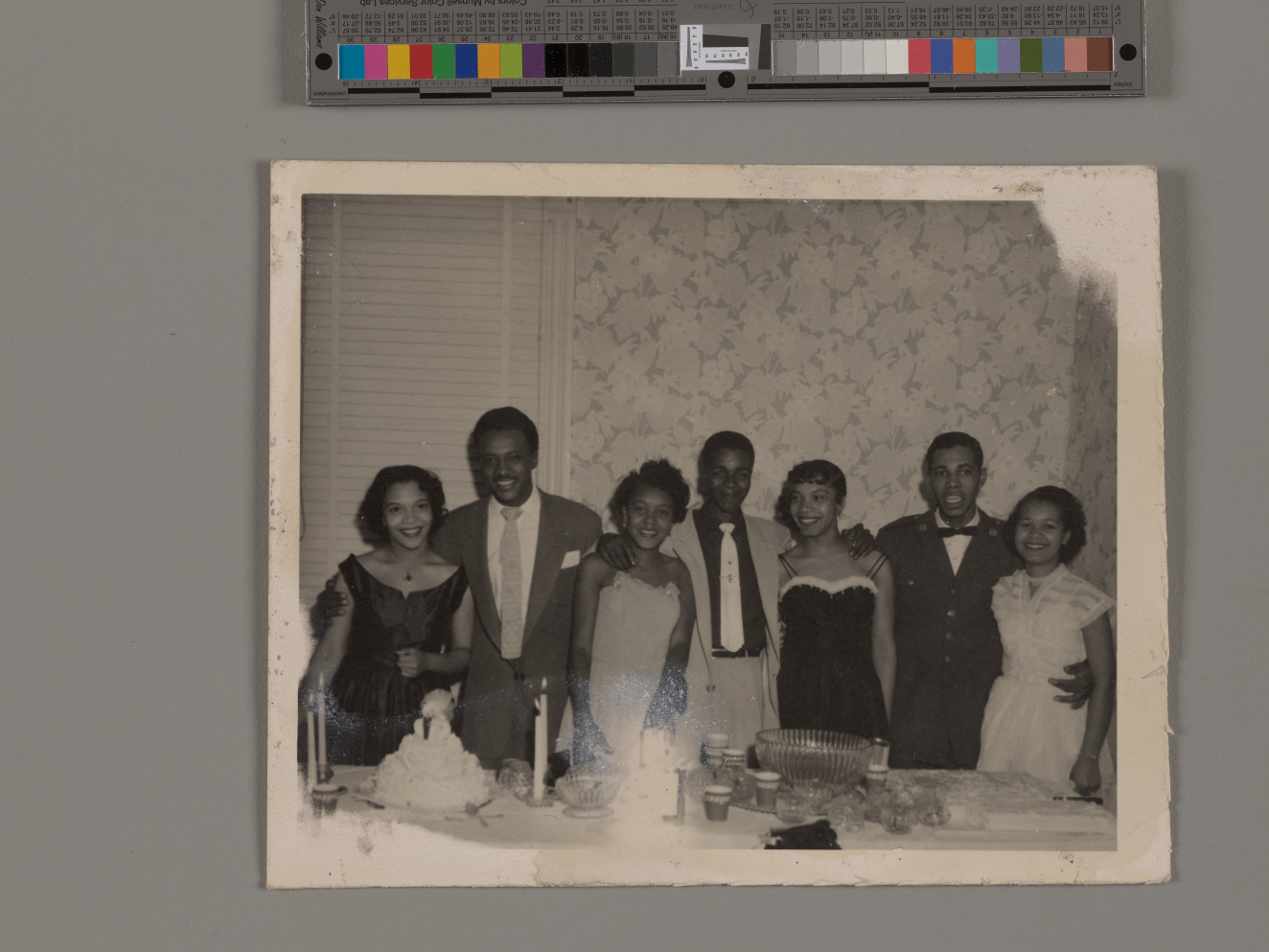 Snapshot of seven dark-skinned people dressed up in suits and cocktail dresses, standing behind a table set with food and drink. They smile at the photographer, arms around each other.