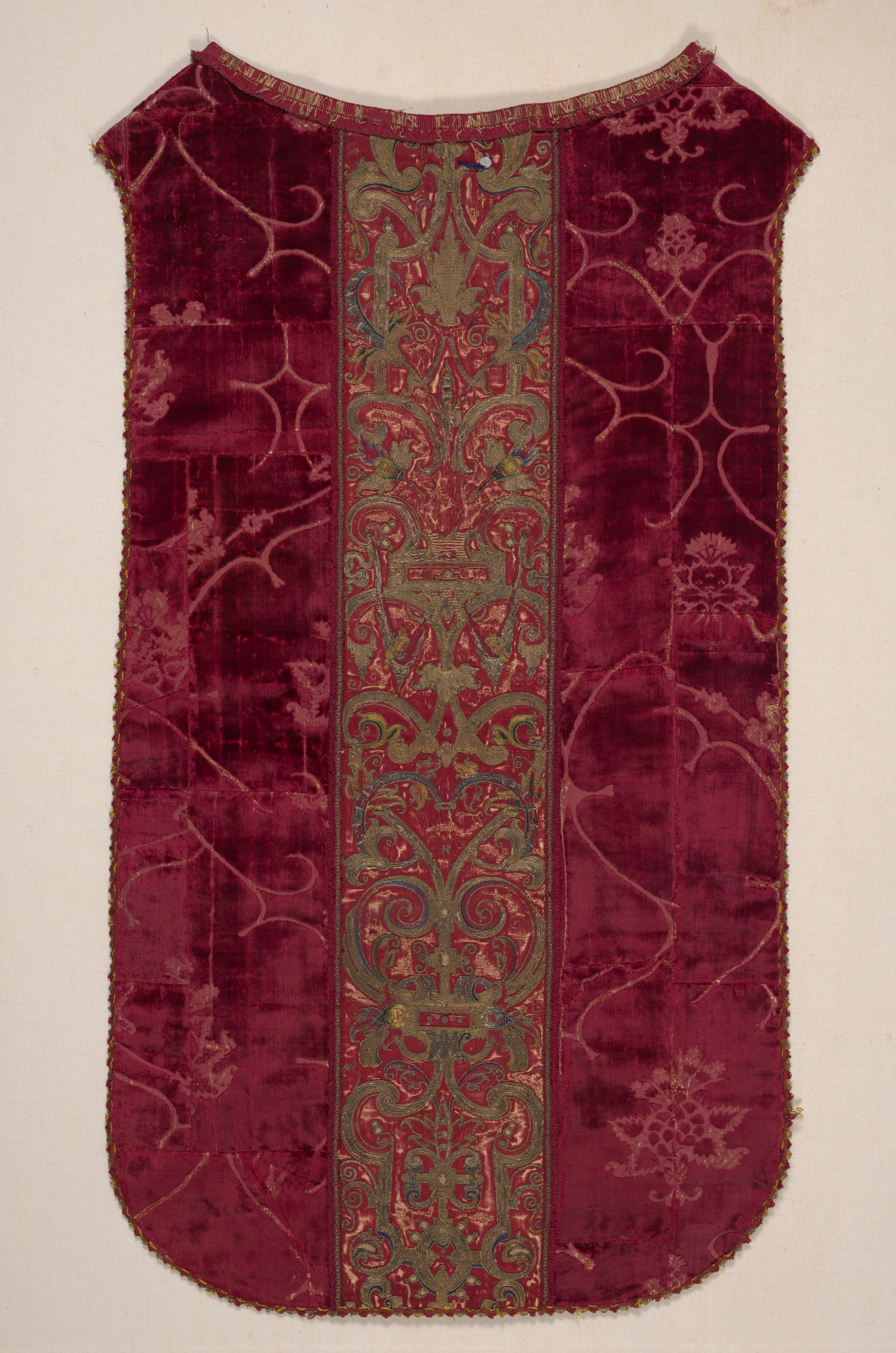 A vest-like garment with a central vertical band of green and gold embroidery showcasing an ornate floral pattern, framed by two panels of plush velvet red with subtle woven design