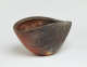 Angled view of a clam-shaped stone bowl with shell-like etchings on its red and brown exterior and a red-orange interior. Its rim dips at the sides forming a wave-like shape. 