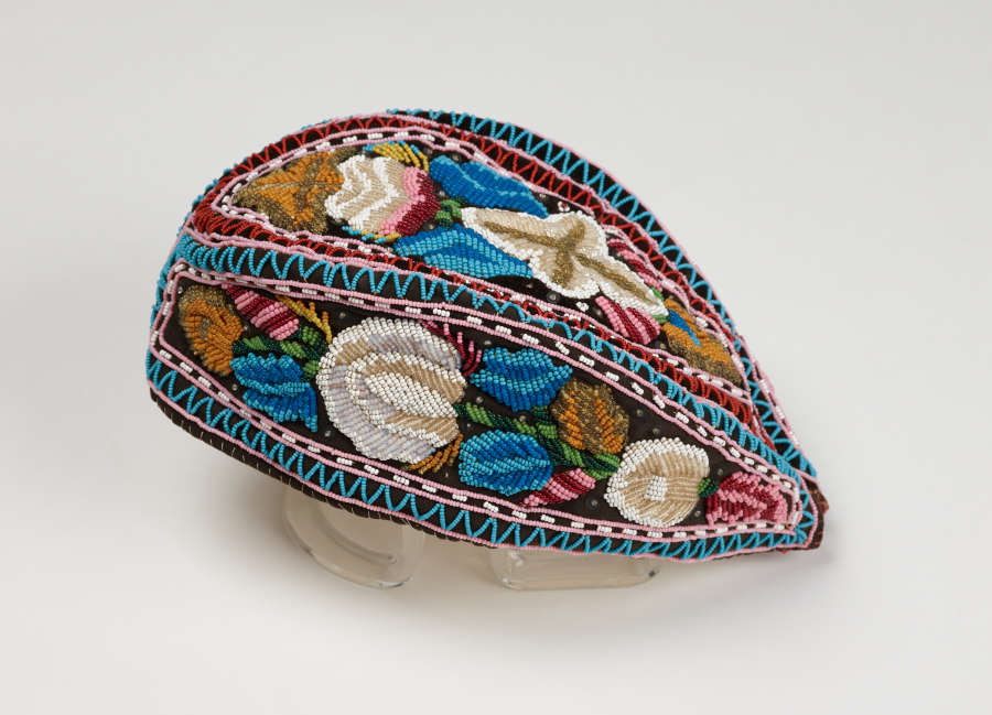 An almond shaped embroidered cap, which features 3 blocks of blue, white, green, and pink floral embroidery which are framed and divided by blue, pink, and white geometric patterns.