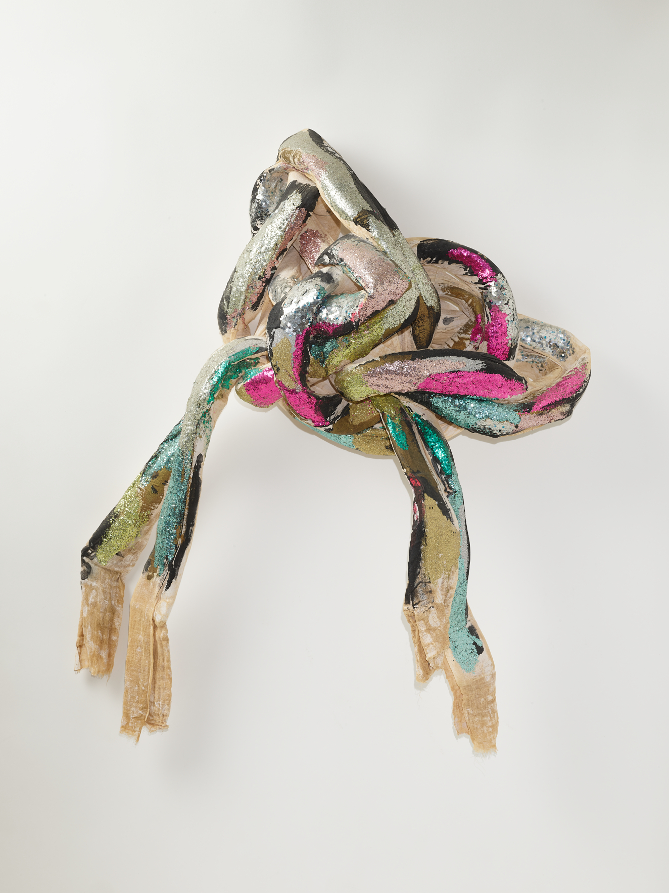 A knotted bundle of textile strips with metallic silver, pink, green, and black cloth, with long frayed ends hanging down loosely. 