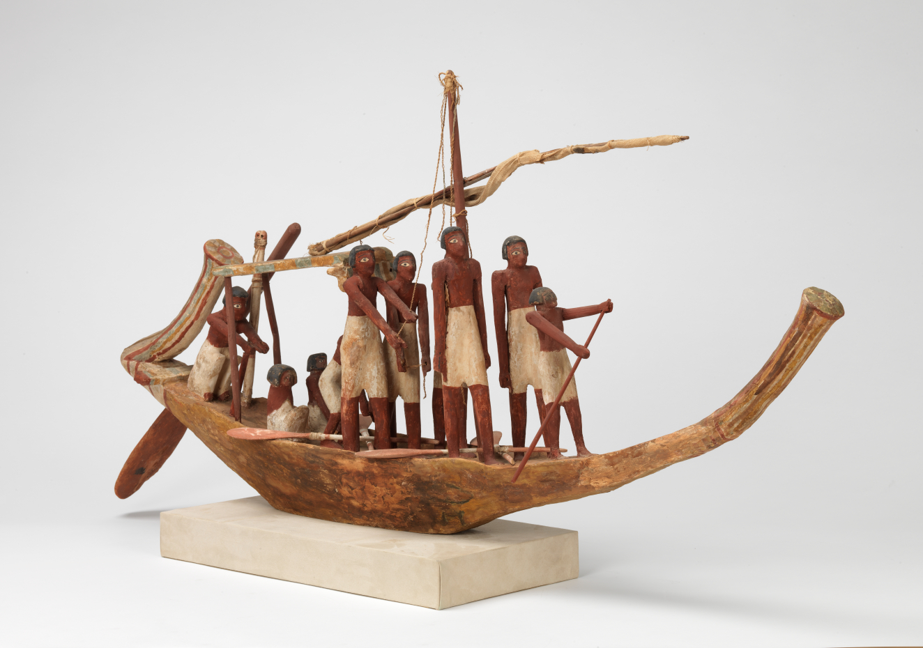 A carved wooden boat model on a rectangular off-white base with a tied up sail manned by sailors with brown skin and white trousers holding oars and ropes.