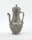 A silver coffee pot with detailed delicate decorations, handle with a lid hinged to it, and spout.