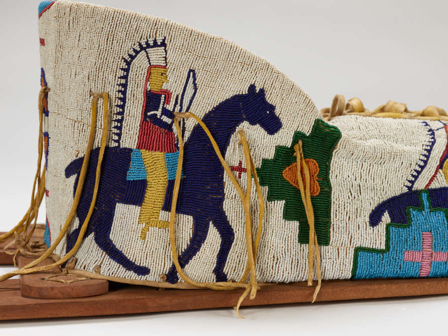 Detail of beaded object with leather ties hanging down and wood supports at bottom. White background beaded with blue horse and colorful rider in headdress, with geometric decoration.  