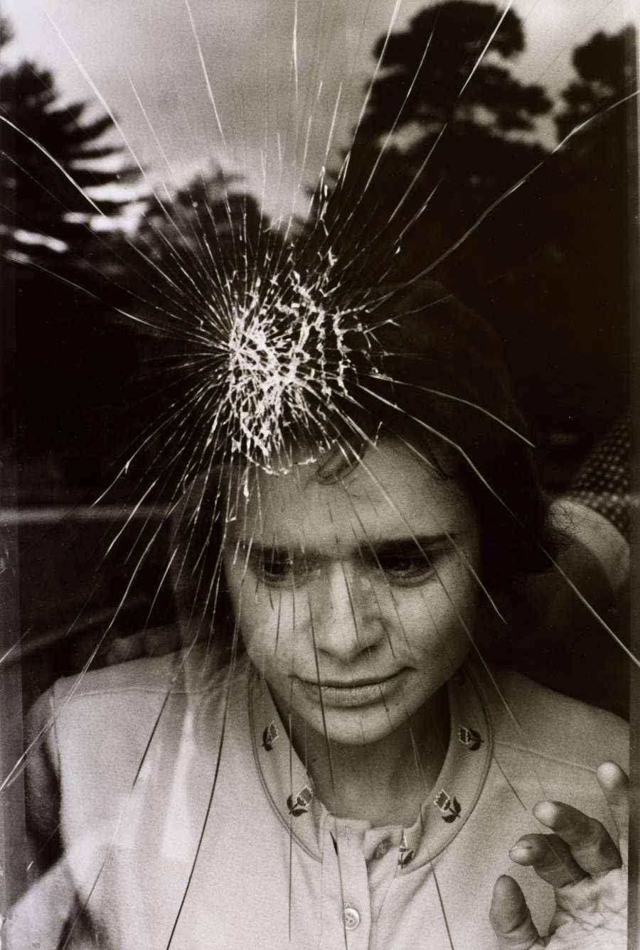 Grayscale photograph of a girl peering out of a cracked window, fingers pressed against the glass and gaze oriented downward. Her face is partially obscured by cracks in the glass.