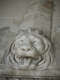 Marble carving depicting a roaring lion’s head with a flowing mane emerging from a horizontal surface, its expression slack-jawed with the eyes rolling back and tongue lolling out.