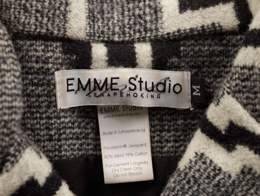 Close-up of the brand and care instructions label on the inside of a gray tweed blazer with black and white geometric patterns. The brand label reads “EMME Studio Lenapehoking.” 
