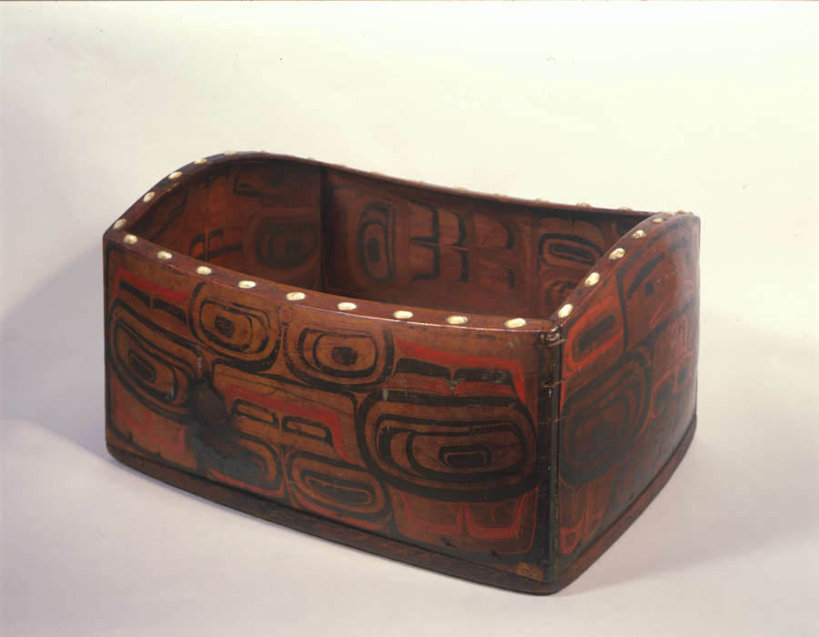 Three-quarter view of a wooden box with red and black graphic motifs resembling creatures and faces. The box’s rim undulates and has a line of large white dots decorating it.