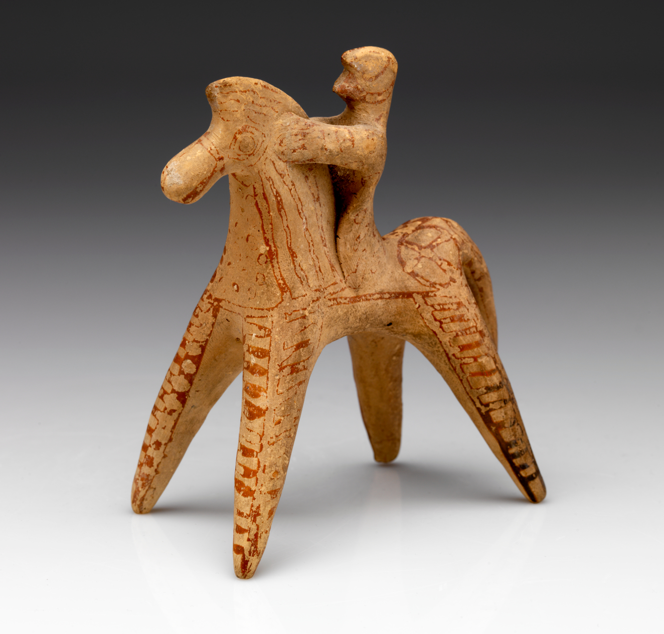 Terracotta figurine featuring the simplified forms of a horse with rider, decorated with slightly faded red painted lines. The painted patterns highlight the horse’s straight legs and figure’s upright posture.
