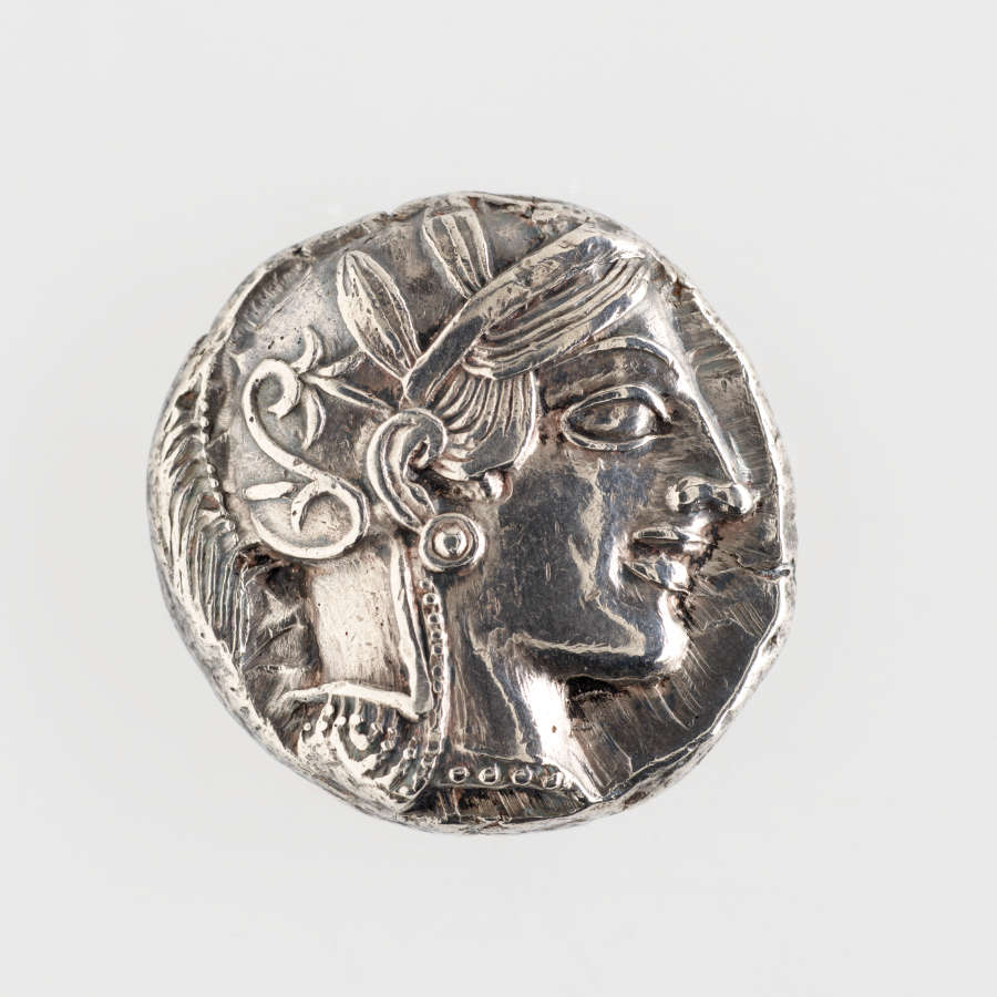 Front of an irregularly round silver coin, embossed with the side profile of a wreathed and ornamented man.