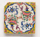 Square wall tile with a chipped upper left corner. It features floral pink, green, and yellow motifs connecting into swirling blue motifs to form a circle. The corners are yellow.