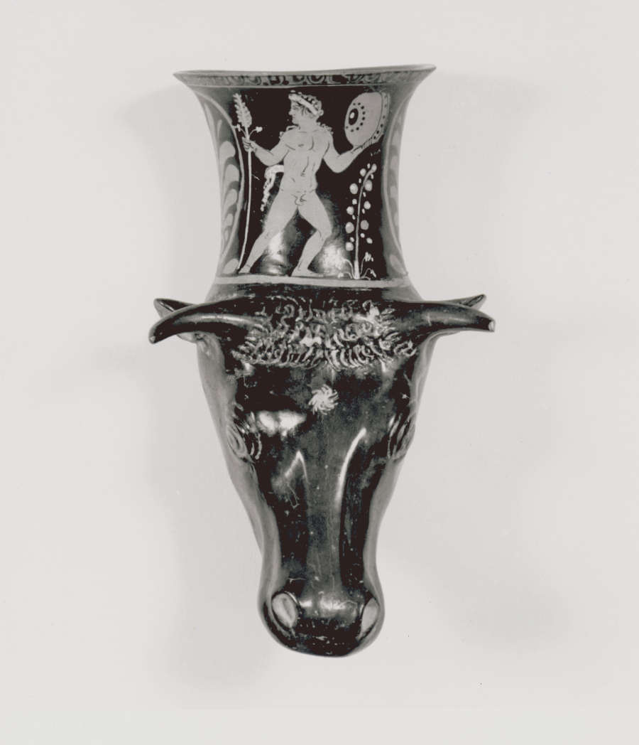 Monochrome photo of a  cup with a sculpted cow’s head as its base, extending into the body of the cup, which is illustrated with men besides large floral motifs. 
