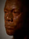 Close-up three quarter view of a detailed, wood-carved face in dim lighting depicting a male figure with afrocentric facial features. The sculpture features nuanced textures, mostly cast in shadow.