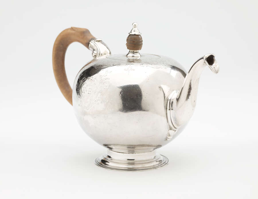 A bulbous silver teapot with a wooden finial and handle connected with decorative silver pieces. There are delicate engraved marks on the body and lid.