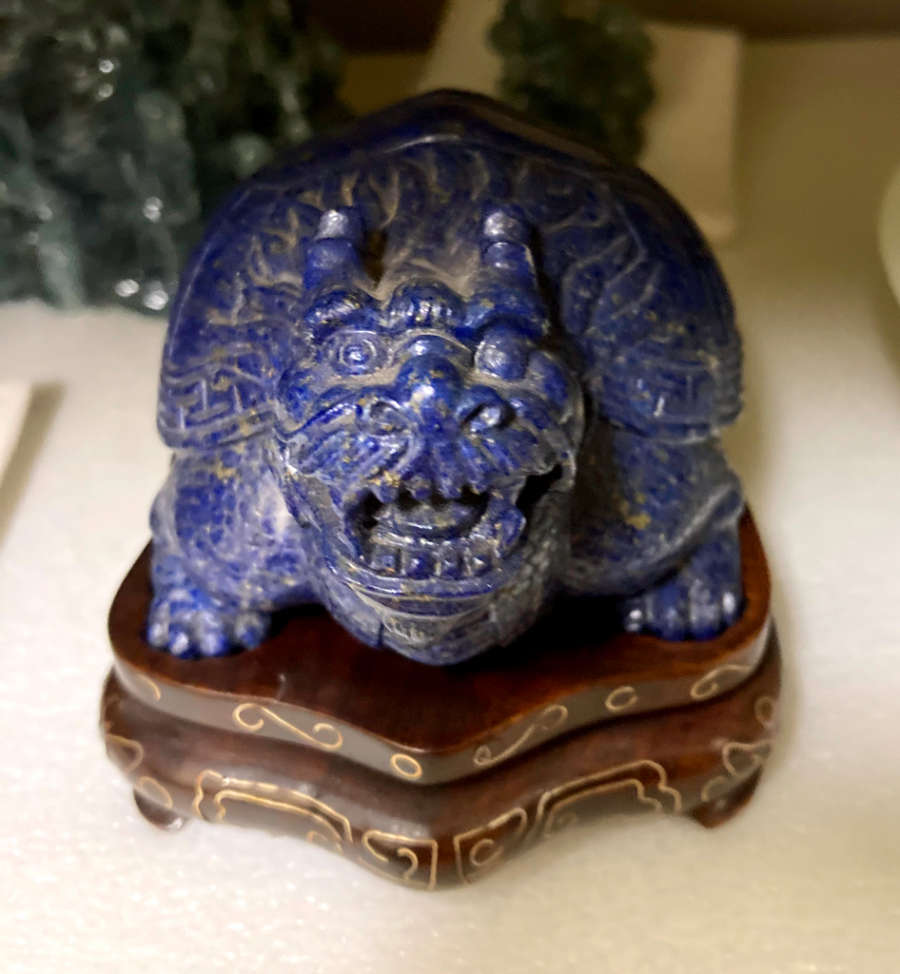 Head-on view of a blue sculpture of a horned tortoise, whose surface is covered with carved geometric patterns. It stands on a brown base with steps and golden patterning. 