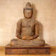 A weathered wooden Buddha statue sitting cross-legged on a wooden platform, with hands resting in the lap, with a serene expression and closed eyes. 