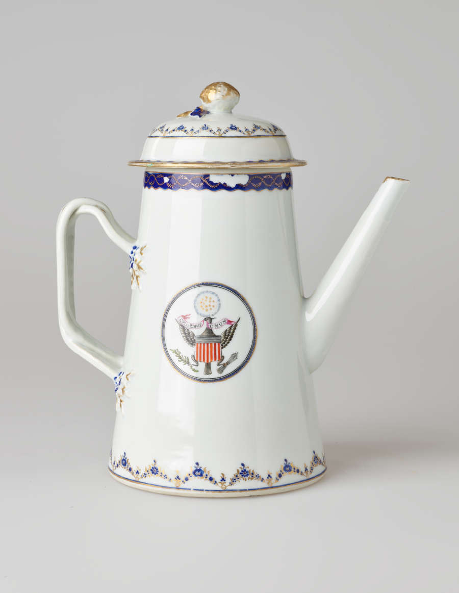A white coffee pot with dark blue and gilded decorations, a straight spout, angular handle, and heraldry imagery in a circle on the body of the coffee pot.