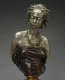Muted bronze bust of a woman gazing downwards with hair in twists, gold earrings, and beaded necklaces accentuating her collarbones above a dark strapless tightly-wrapped top mounted on a gold base.
