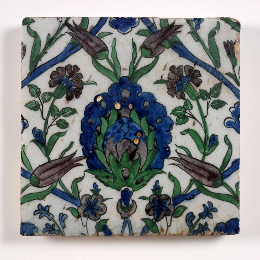 White square tile painted with a dense symmetrical floral pattern in blues, blacks and greens. A large maned flower occupies the center, with smaller flowers of varying shapes surrounding it.