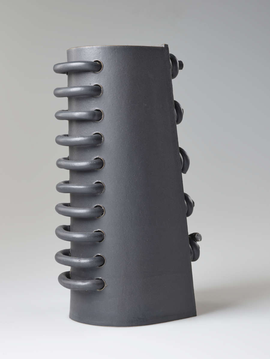 Diagonal back view of a gray industrial-like cylinder with one flat periphery. Ten large vertically aligned loops are threaded through and protruding from the rounded back side
