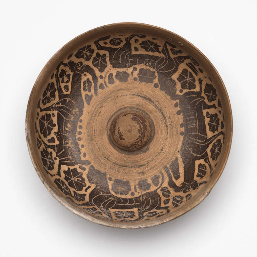 Top-view of the worn brown ceramic bowl with black patterning of animals walking along its rim surrounded by floral motifs. A small dome rises from the bowl's wide, unpainted center.