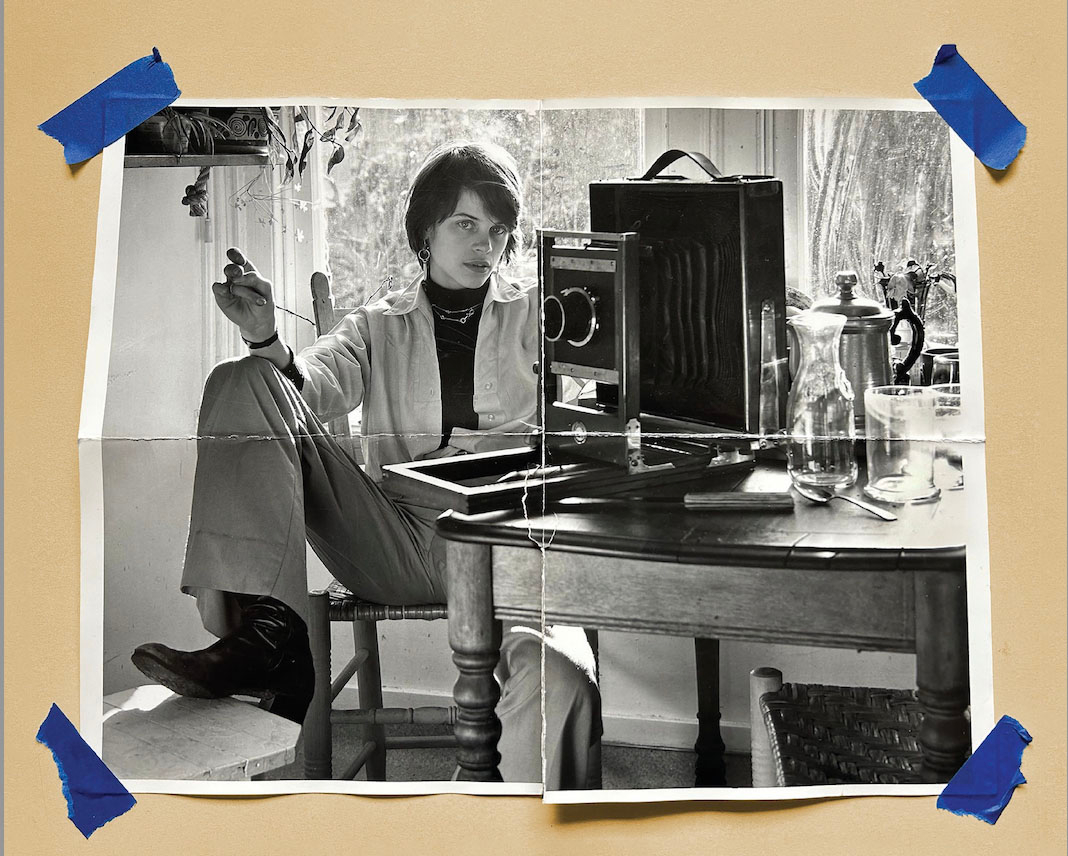 Folded photograph taped to a wall, showing a woman sitting on a chair in front of a window. A large format camera and drinking glasses are on the table next to her, and a house plant hangs above her. She has short hair and is wearing boots, and one of her feet is propped up on the chair in front of her.