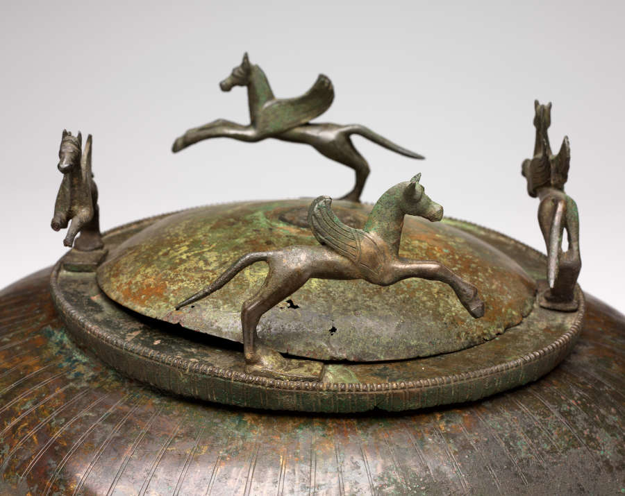 Detail of the bowl’s bronze corroded lid resting on its rim, bringing into focus the engraved marks running down the surface, and all four of the bronze winged horses. 
