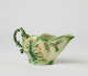  A short boat shaped pitcher green, cream, and brown in color with a sculptural handle and floral decorations.
