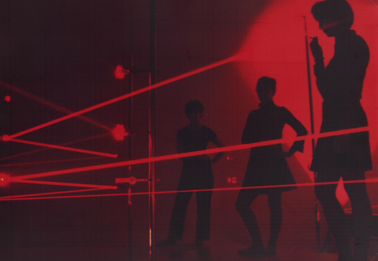 Dark silhouetted figures pose against an immersive glowing red space, intersected by criss-crossing laser beams. 
