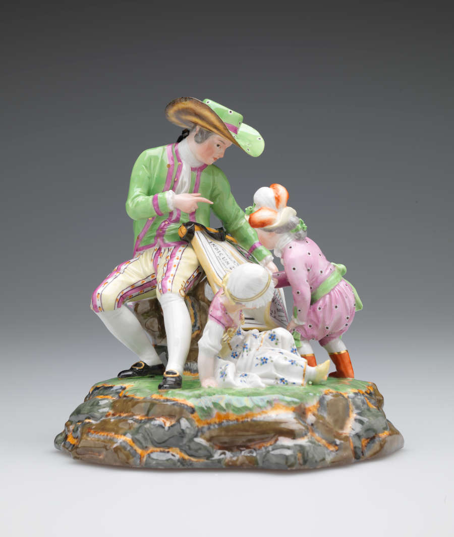 A sculpture with three figures in elaborate historical clothing. A seated light-skinned figure holds a roll of maps while one child examines. A second child sits near them.