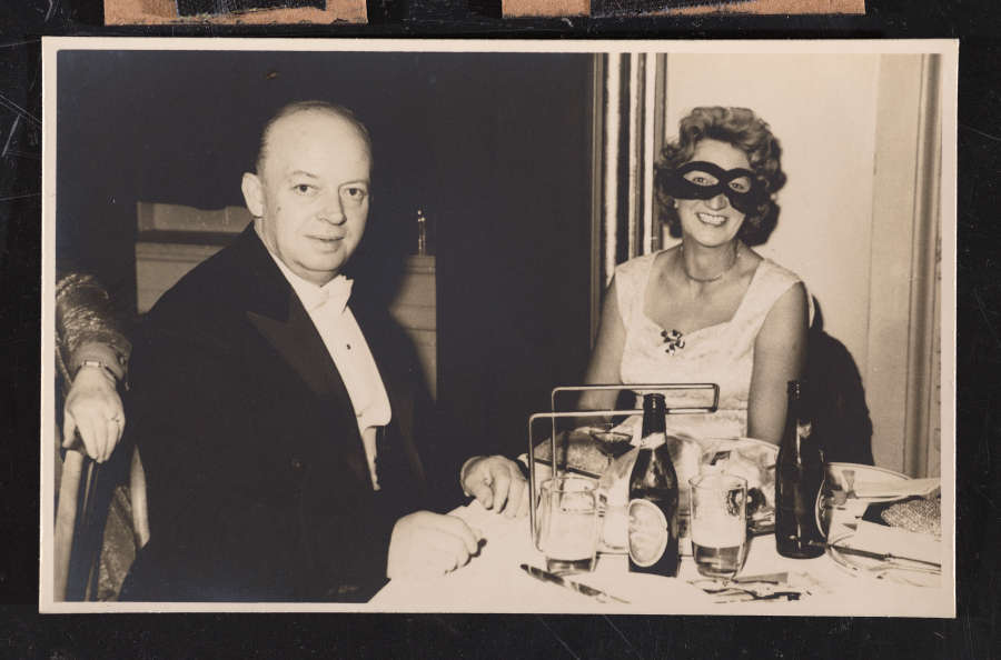Snapshot of two smiling older light-skinned people sitting at a table. He is bald and wears a tuxedo. She wears a white dress and a black mask over her eyes. 