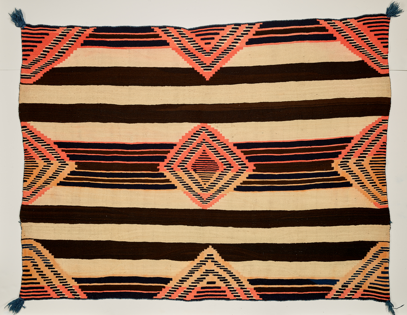 Black and cream striped woven blanket with concentric salmon pink and yellow diamonds overtop. The diamond pattern is cut off along the edges and dark green tassels adorn each corner.