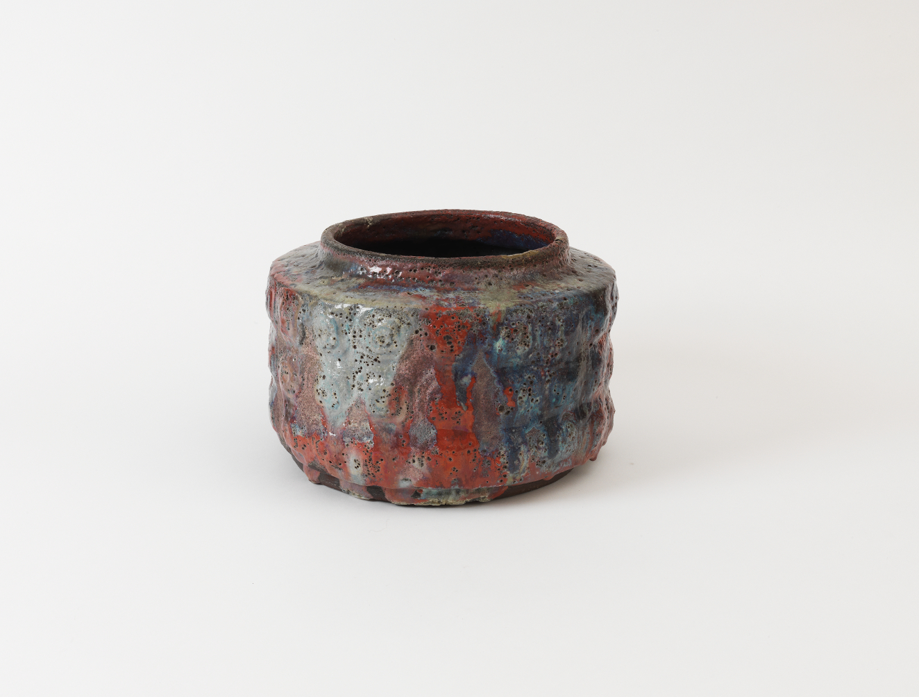 Small, cylindrical ceramic jar glazed with a glossy finish with a rough, ribbed surface covered in pores, the earthy red clay textured with patches of blue and gray tones.