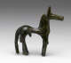 Side-view of a bronze horse figurine with a green patina, and an exaggeratedly thin torso, thick neck, long legs and tail and long narrow snout.