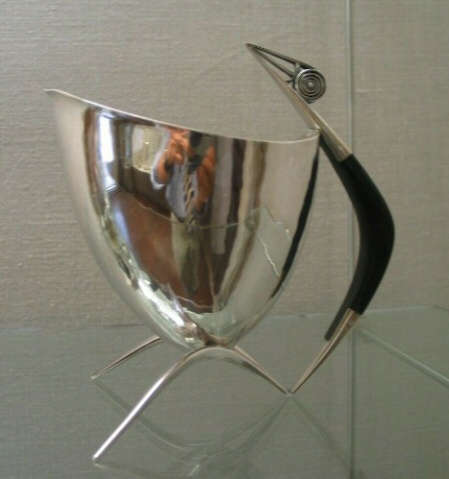 Sleek silver vessel with a curved body atop three pointed legs, featuring a black handle with a spiral detail and sharp ends which stretch above the vessel's top.