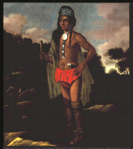 A brown skinned man wears a beaded headdress and necklace, with a red cloth around his waist. He carries a wooden staff with a knife tucked at his side.