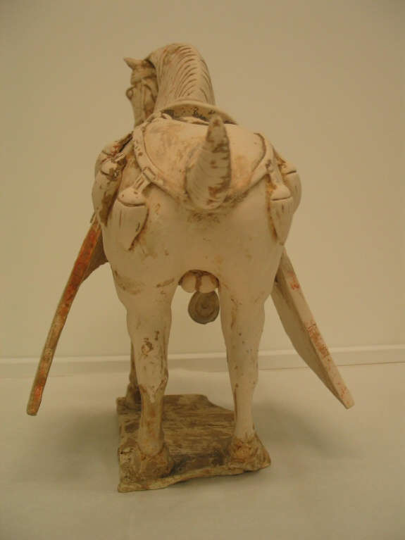 Back-view of a ceramic sculpture of a saddled horse, with a wound tail and thick mane, standing on a thin base. Visible are its ornamental belts and hanging saddle blanket.