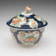 A lidded sugar bowl with floral decorations in gilded vignettes, surrounded by dark blue glaze, the finial on the lid is a sculptural red, white, and yellow flower.