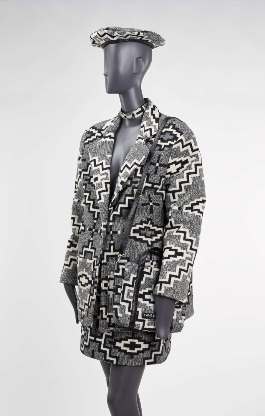 Three-quarter view of a mannequin wearing a gray tweed blazer, with a matching hat, mid-sized purse, skirt and choker. The set features bold black and white geometric designs throughout. 