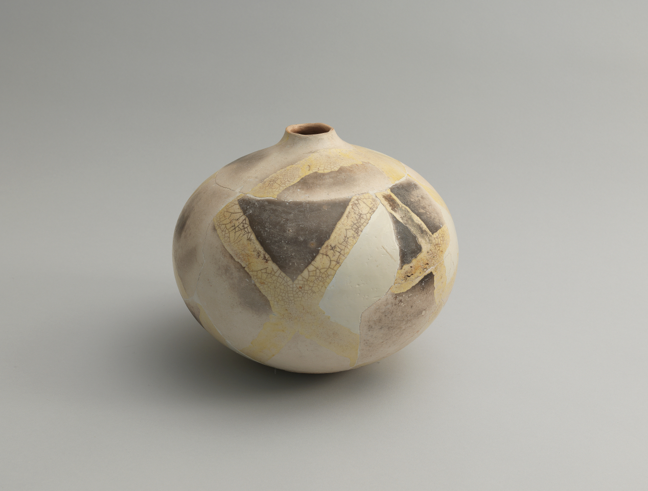 A spherical vessel with a narrow opening, patterned with muted beige, gray, and cream patches intersected by faded gold bands that create an abstract geometric pattern.
