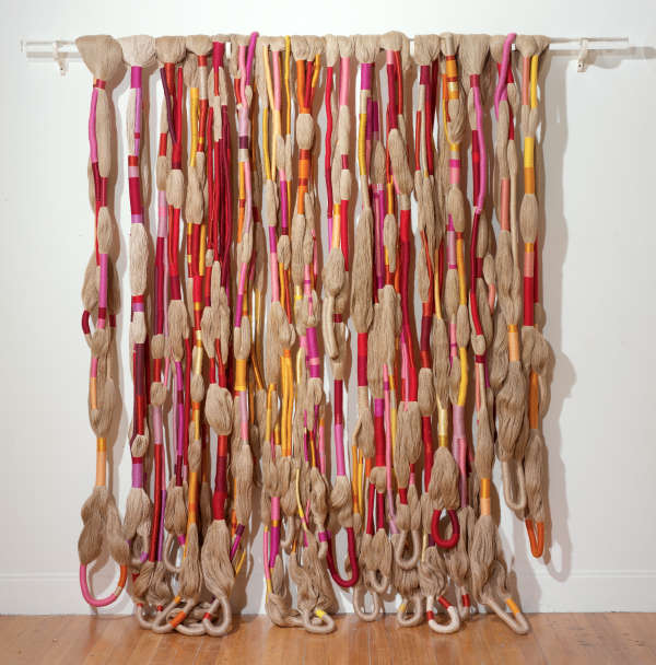 Textural installation featuring long bunches of beige yarn, tied in intervals with pink, red, and orange ribbons, hanging from a horizontal pole propped on a wall, creating a curtain-like display.