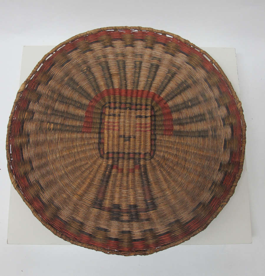 Circular basket, woven in a radial pattern with a bordered edge, in undyed, green, and red straw-like fibers. Woven in is an image of a necklaced head dressed human.