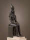 A dark metal statue of a human figure seated on a throne with a falcon head, wearing a tall, cone-like crown with a cobra.