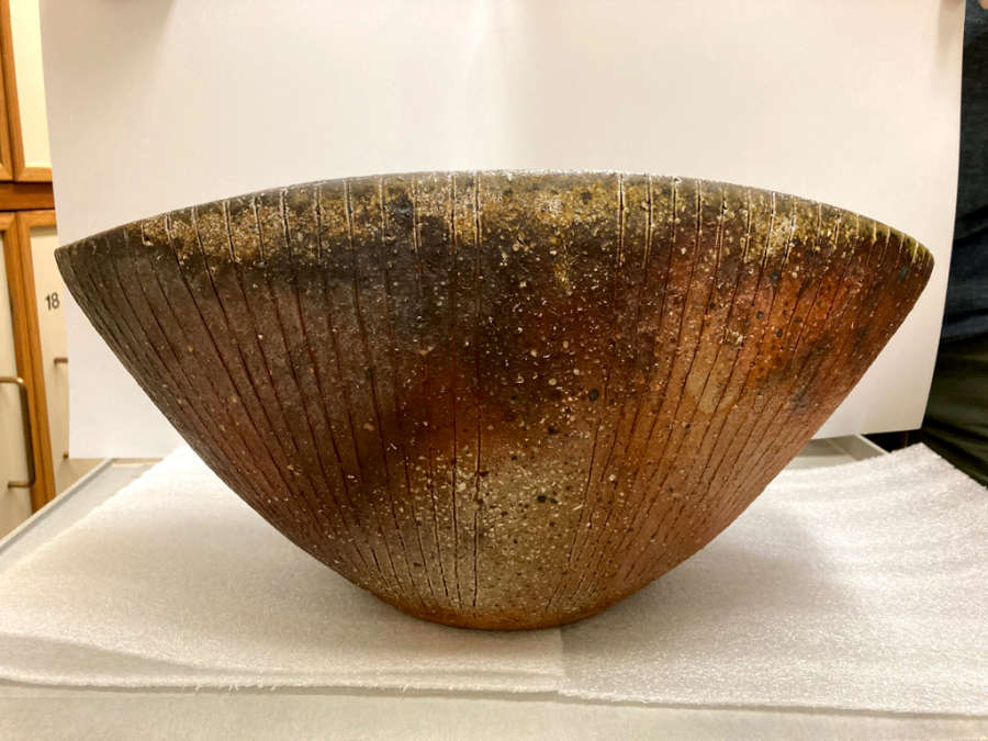 View in storage of a clam-shaped bowl with shell-like etchings. Its rim is speckled with yellow earthy tones fading into the orange-brown splotches of its body.