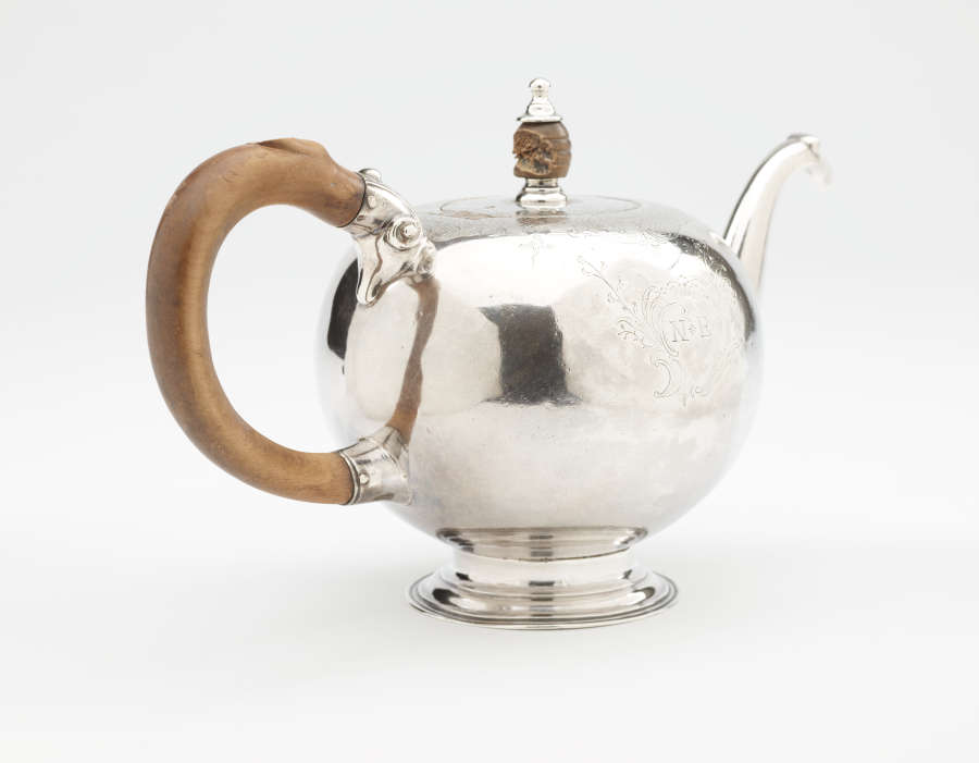 A bulbous silver teapot with a wooden finial and handle connected with decorative silver pieces. There are delicate engraved marks on the body and lid.