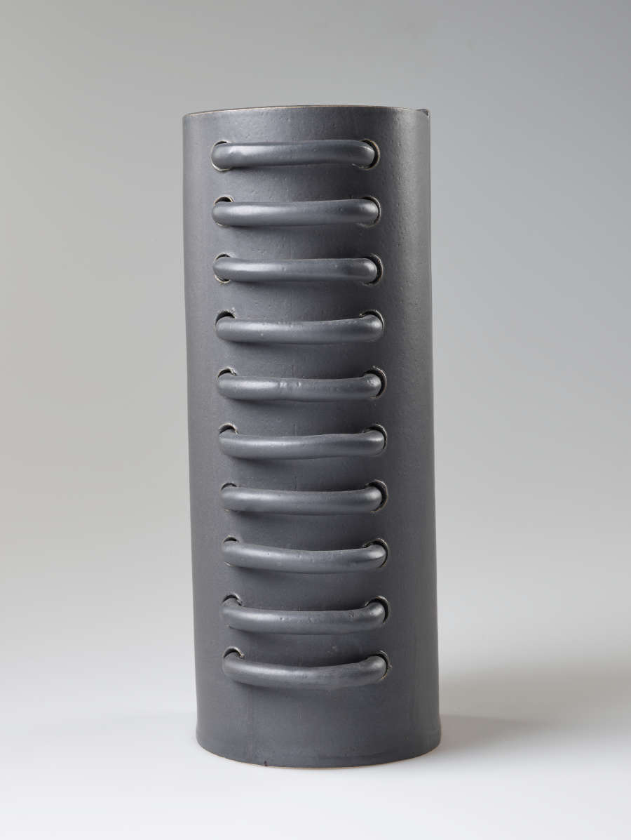  Back view of a gray industrial-like ceramic cylinder with ten large vertically aligned loops threaded through and protruding from the center.
