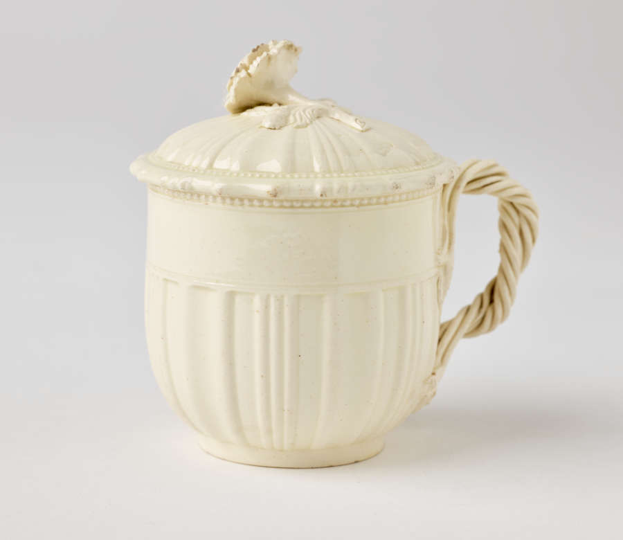  A cream-colored cup with a sculptural swirled handle, and lid with sculpted finial.