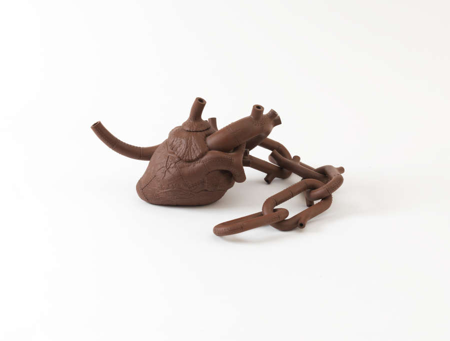Brown ceramic teapot shaped like an anatomical human heart with fine vein-like textures on the surface and extended artery-like spouts, accompanied by a large chain link curling to the side.